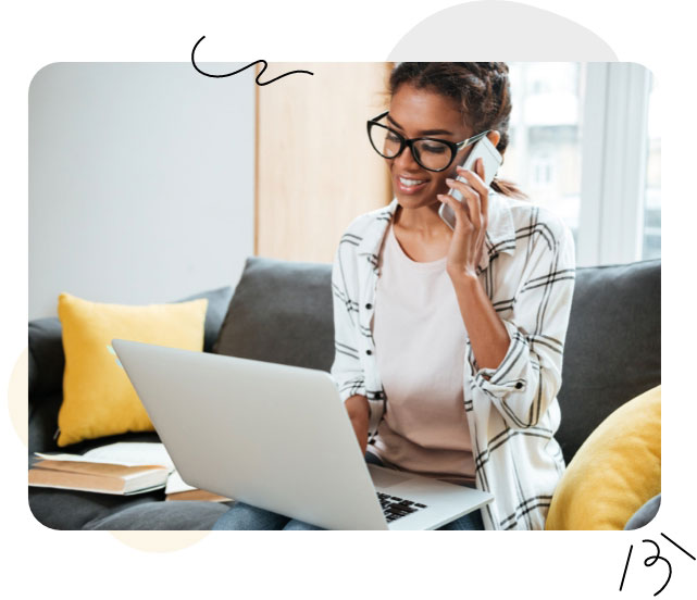 Una mujer con gafas está sentada en un sofá, con un portátil y un teléfono en las manos, realizando varias tareas a la vez con eficacia.